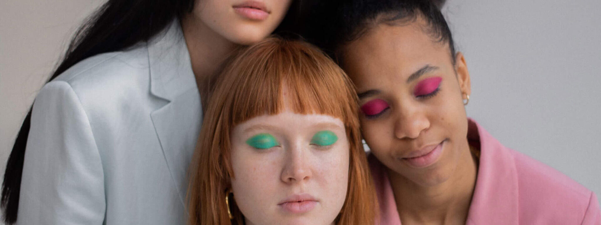 Drei Frauen in modischen Anzügen mit farbigem Lidschatten in Pastelltönen, die eine entspannte Pose einnehmen.