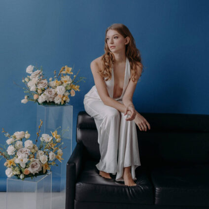 Braut in einem eleganten Kleid, sitzend auf einem schwarzen Sofa vor einem blauen Hintergrund, mit Blumengestecken auf transparenten Podesten daneben.